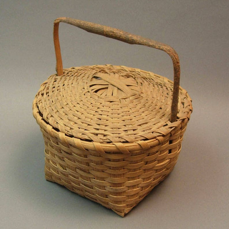 May include: A small, round, woven basket with a lid and a wooden handle. The basket is made of natural materials and has a light brown colour.