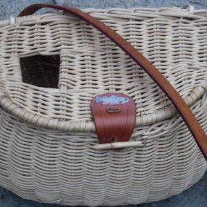 May include: A woven wicker fishing creel with a brown leather strap and a small fish design on the front.