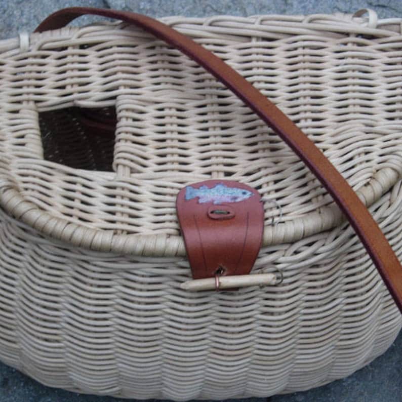 May include: A woven wicker fishing creel with a brown leather strap and a small fish design on the front.