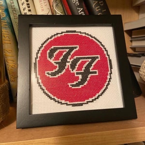 May include: Framed cross-stitch art featuring the Foo Fighters logo. The design includes a red circle with the band's initials, "FF," in black. The artwork is displayed in a black frame and rests on a wooden surface.