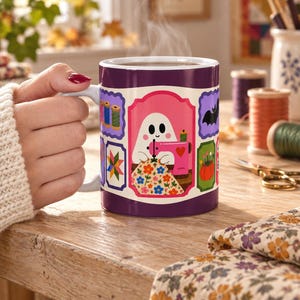 May include: A purple ceramic mug featuring a central illustration of a white ghost using a pink sewing machine to stitch a floral fabric. The design includes smaller rectangular frames displaying spools of thread, a quilt block, a bat, and a pumpkin.