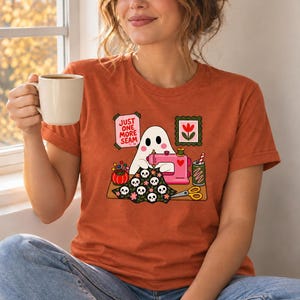 Woman holding a mug, wearing an Autumn (burnt orange) t-shirt with the "Just One More Seam" folk-art ghost sewing print. Color: Autumn.