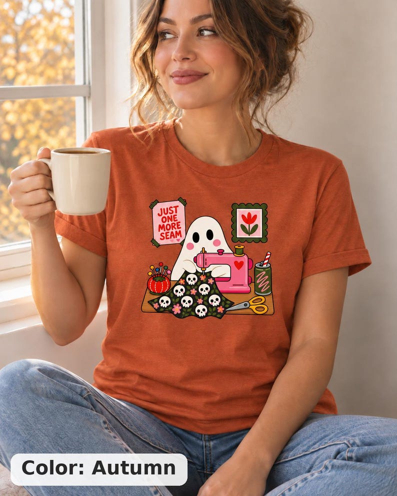 Woman holding a mug, wearing an Autumn (burnt orange) t-shirt with the "Just One More Seam" folk-art ghost sewing print. Color: Autumn.