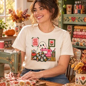 Woman smiling at a sewing table in a bright sewing room, wearing a natural-colored t-shirt with the "Just One More Seam" folk-art ghost sewing at a pink sewing machine.