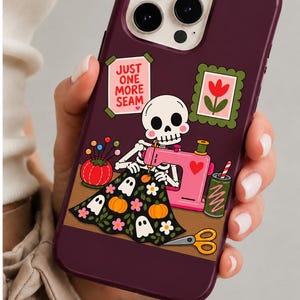 Könnte beinhalten: Eine dunkelviolette Smartphone-Hülle mit einer Grafik eines Skeletts, das an einer rosa Nähmaschine näht. Das Design zeigt ein Nadelkissen, eine Schere, ein grünes Getränk, eine gerahmte Blume und ein Schild mit der Aufschrift JUST ONE MORE SEAM über schwarzem Stoff mit Kürbissen und Geistern.