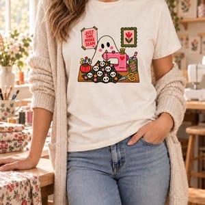 Woman in a cream cardigan and jeans wearing a natural-colored t-shirt printed with the "Just One More Seam" folk-art ghost sewing at a pink sewing machine, standing in a cozy sewing room.