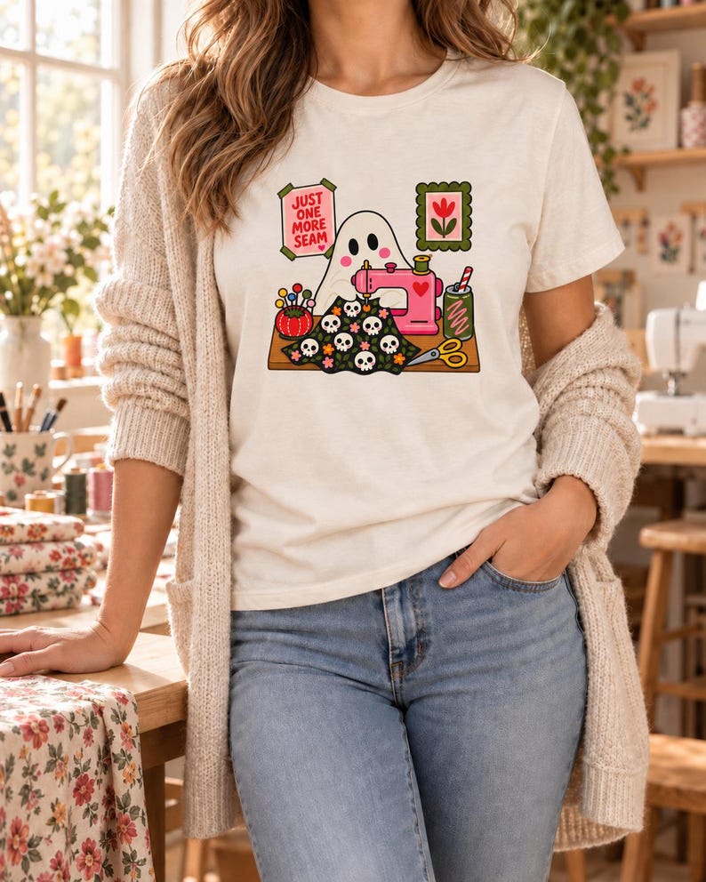 Woman in a cream cardigan and jeans wearing a natural-colored t-shirt printed with the "Just One More Seam" folk-art ghost sewing at a pink sewing machine, standing in a cozy sewing room.