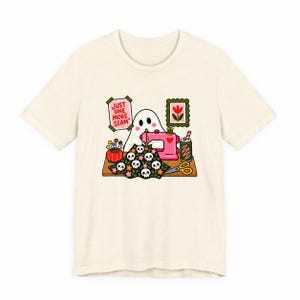 Plain flat product photo of a natural-colored t-shirt with the "Just One More Seam" folk-art ghost sewing print, showing what ships.