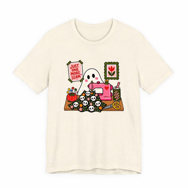 Plain flat product photo of a natural-colored t-shirt with the "Just One More Seam" folk-art ghost sewing print, showing what ships.