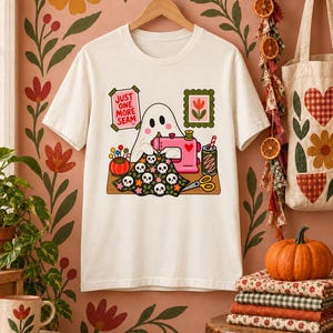 Natural-colored t-shirt with the "Just One More Seam" folk-art ghost sewing print hanging on a styled wall with a fall floral tote and pumpkins.