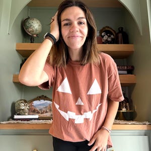 Könnte beinhalten: Ein rostfarbenes Kurzarm-T-Shirt mit einem großen weißen Aufdruck eines Kürbisgesichts auf der Vorderseite, bestehend aus zwei dreieckigen Augen, einer kleinen dreieckigen Nase und einem gezackten, zahnigen Grinsen.