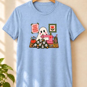 Light Blue t-shirt with the "Just One More Seam" folk-art ghost sewing print, hanging against a plain wall. Color: Light Blue.