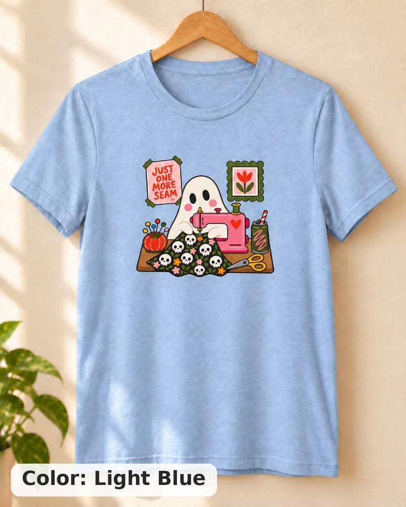 Light Blue t-shirt with the "Just One More Seam" folk-art ghost sewing print, hanging against a plain wall. Color: Light Blue.