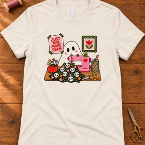 Natural t-shirt with the "Just One More Seam" folk-art ghost sewing print, laid flat on a wooden table. Color: Natural.