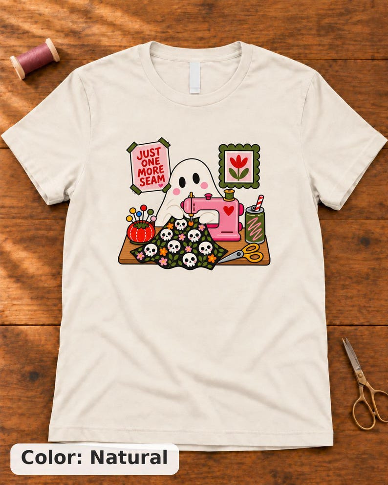 Natural t-shirt with the "Just One More Seam" folk-art ghost sewing print, laid flat on a wooden table. Color: Natural.