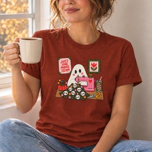 Woman holding a mug, wearing a Rust (deep red-brown) t-shirt with the "Just One More Seam" folk-art ghost sewing print. Color: Rust.