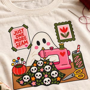 Close-up of the printed "Just One More Seam" folk-art ghost design on a natural-colored t-shirt, showing the ghost, pink sewing machine, skull-patterned quilt and flower frames.