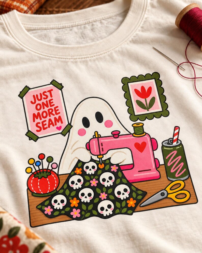 Close-up of the printed "Just One More Seam" folk-art ghost design on a natural-colored t-shirt, showing the ghost, pink sewing machine, skull-patterned quilt and flower frames.