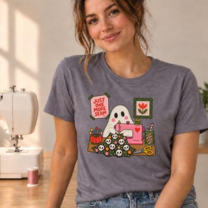 Woman in a sewing room wearing a Storm (slate blue-gray) t-shirt with the "Just One More Seam" folk-art ghost sewing print. Color: Storm.