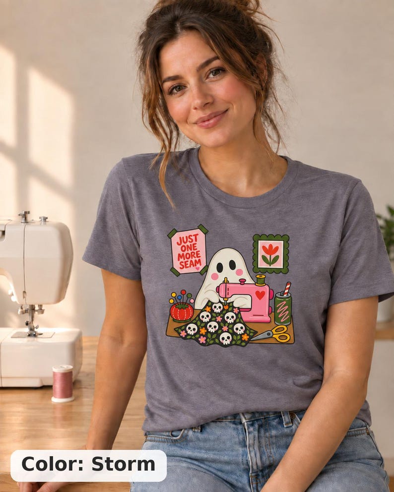 Woman in a sewing room wearing a Storm (slate blue-gray) t-shirt with the "Just One More Seam" folk-art ghost sewing print. Color: Storm.