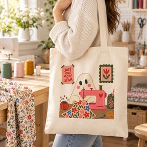 May include: A cream-colored canvas tote bag featuring a graphic illustration of a ghost using a pink sewing machine. The design includes a red pincushion, floral fabric, scissors, a drink can, and a sign that reads Just One More Seam.