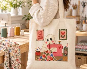 May include: A cream-colored canvas tote bag featuring a graphic illustration of a ghost using a pink sewing machine. The design includes a red pincushion, floral fabric, scissors, a drink can, and a sign that reads Just One More Seam.