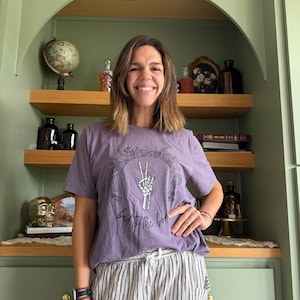 Könnte beinhalten: Ein kurzärmeliges T-Shirt in meliertem Lila mit einem schwarzen Grafikdruck einer Skeletthand, die ein Friedenszeichen formt, umgeben von einem Blumenkranz, mit dem Text "bad to the bone" in Schreibschrift unter der Grafik.