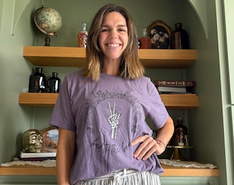 May include: A heathered purple short-sleeve t-shirt featuring a black graphic print of a skeleton hand making a peace sign surrounded by a floral wreath, with the text "bad to the bone" written in a cursive script underneath the graphic.