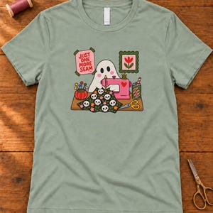 Sage (muted green) t-shirt with the "Just One More Seam" folk-art ghost sewing print, laid flat on a wooden table. Color: Sage.