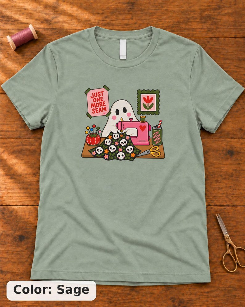 Sage (muted green) t-shirt with the "Just One More Seam" folk-art ghost sewing print, laid flat on a wooden table. Color: Sage.
