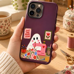 Könnte beinhalten: Eine dunkelviolette Smartphone-Hülle mit einer Grafik eines weißen Geistes, der an einer rosa Nähmaschine näht. Das Design zeigt ein geblümtes Stoffstück, ein rotes Nadelkissen, ein grünes Getränk und ein Schild mit der Aufschrift JUST ONE MORE SEAM.