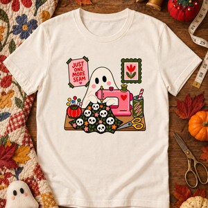 Natural-colored t-shirt with the "Just One More Seam" folk-art ghost sewing at a pink sewing machine printed on the front, laid flat on a wooden table with autumn leaves, pumpkins and sewing notions.