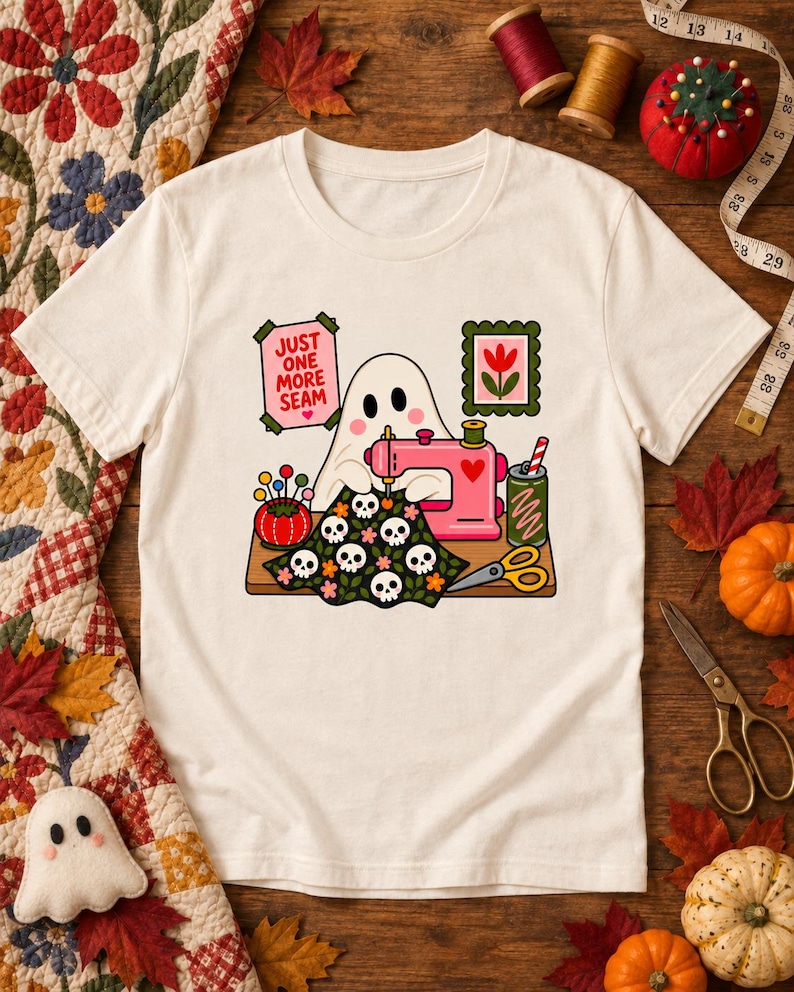 Natural-colored t-shirt with the "Just One More Seam" folk-art ghost sewing at a pink sewing machine printed on the front, laid flat on a wooden table with autumn leaves, pumpkins and sewing notions.