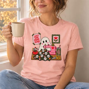 Woman holding a mug, wearing a Peach (soft pink) t-shirt with the "Just One More Seam" folk-art ghost sewing print. Color: Peach.