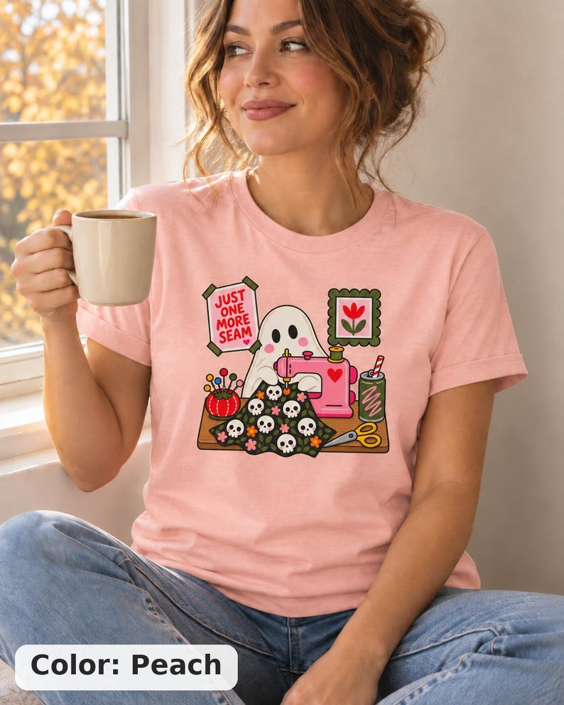 Woman holding a mug, wearing a Peach (soft pink) t-shirt with the "Just One More Seam" folk-art ghost sewing print. Color: Peach.
