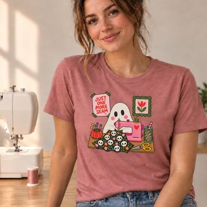 Woman smiling, wearing a Mauve (dusty rose) t-shirt with the "Just One More Seam" folk-art ghost sewing print. Color: Mauve.