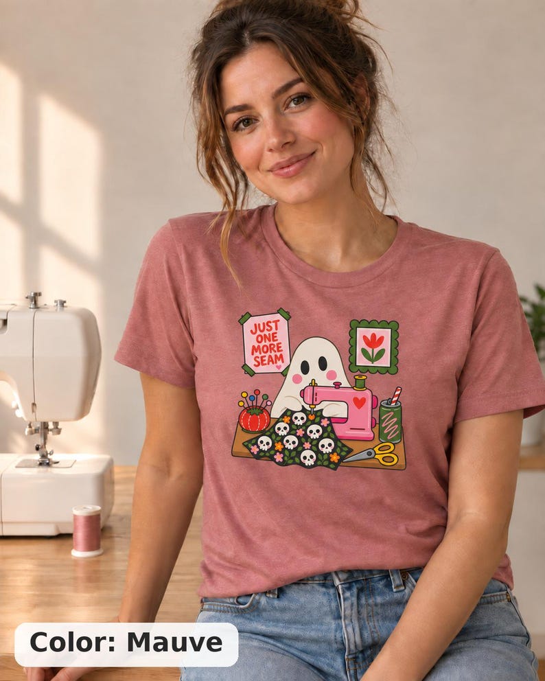 Woman smiling, wearing a Mauve (dusty rose) t-shirt with the "Just One More Seam" folk-art ghost sewing print. Color: Mauve.