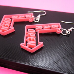 May include: A pair of pink open sign earrings with silver hooks. The earrings are shaped like arrows pointing to the right, with the word "OPEN" written on them.