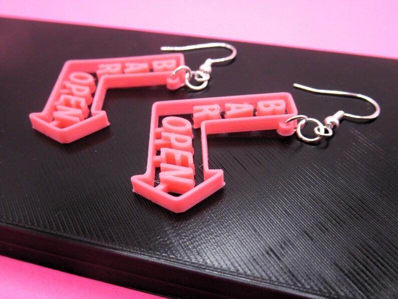 May include: A pair of pink open sign earrings with silver hooks. The earrings are shaped like arrows pointing to the right, with the word "OPEN" written on them.