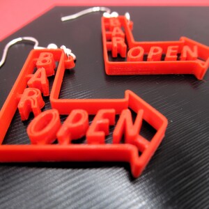 May include: A pair of red 3D printed earrings shaped like arrows with the words "BAR OPEN" on them.