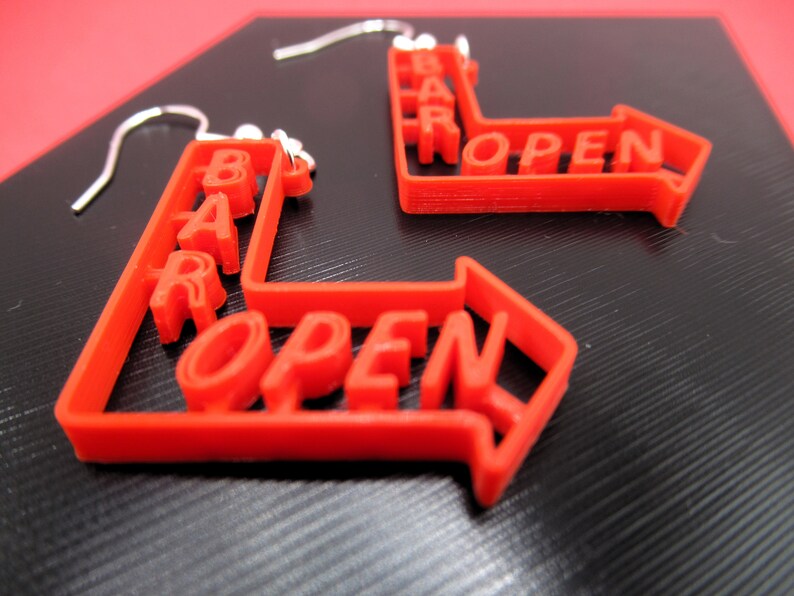 May include: A pair of red 3D printed earrings shaped like arrows with the words "BAR OPEN" on them.