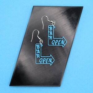 May include: A pair of blue earrings shaped like a bar sign with the words "BAR OPEN" on them. The earrings are on a black background.