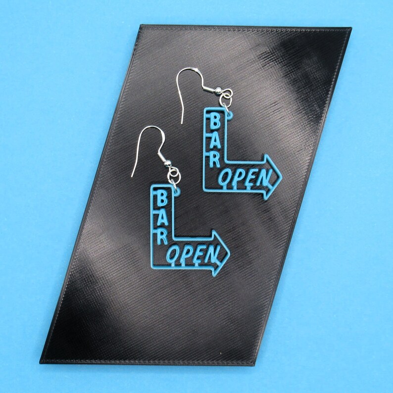 May include: A pair of blue earrings shaped like a bar sign with the words "BAR OPEN" on them. The earrings are on a black background.