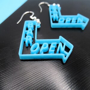 May include: A pair of blue earrings shaped like a directional arrow with the words "BAR OPEN" written on them.