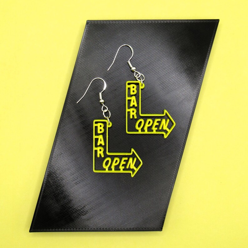 May include: A pair of yellow acrylic earrings shaped like bar signs with the words "BAR OPEN" on them. The earrings are hanging from silver hooks.