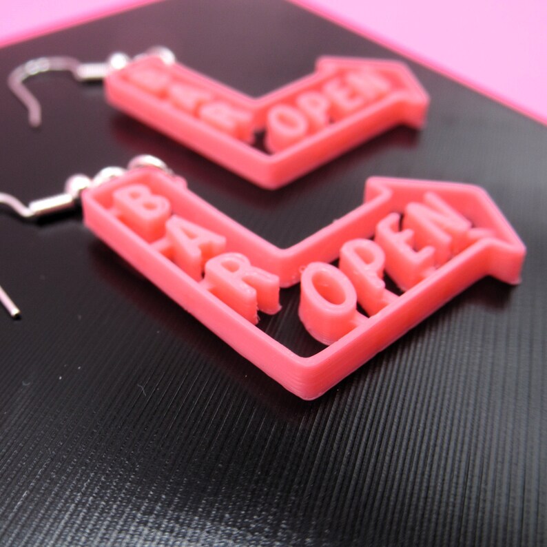 May include: A pair of pink plastic earrings shaped like arrows with the words "Bar Open" on them.