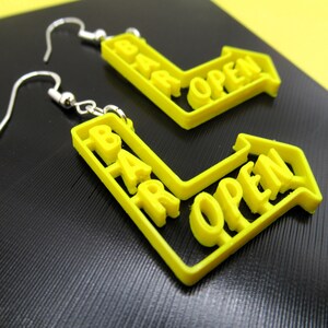 May include: A pair of yellow earrings shaped like a bar sign with the words "BAR OPEN" on them. The earrings are made of plastic and have silver hooks.