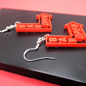 May include: A pair of red earrings shaped like arrows with the words "BAR OPEN" written on them. The earrings are made of plastic and have silver hooks.
