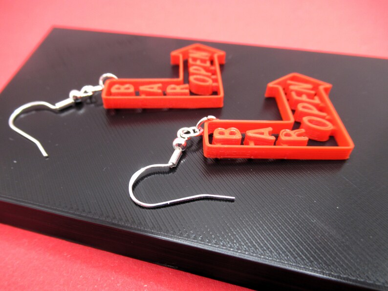 May include: A pair of red earrings shaped like arrows with the words "BAR OPEN" written on them. The earrings are made of plastic and have silver hooks.