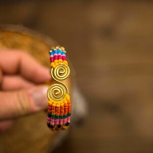 May include: A colorful woven bracelet with a gold metal double spiral design. The bracelet is made with a variety of colors, including red, orange, yellow, green, blue, and pink.